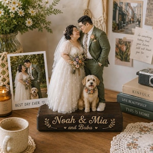 May include: A custom wedding cake topper featuring a miniature bride in a white floral dress, a groom in a green suit, and a small dog. They stand on a dark wooden base engraved with the text Noah & Mia and Bubu, decorated with leaf motifs.