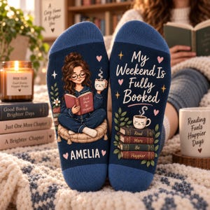 Può includere: Un paio di calzini blu scuro con la grafica di una persona che legge su un cuscino, una tazza a forma di gatto e il testo "My Weekend Is Fully Booked" e "AMELIA". Il design include pile di libri, piccoli cuori e stelle.