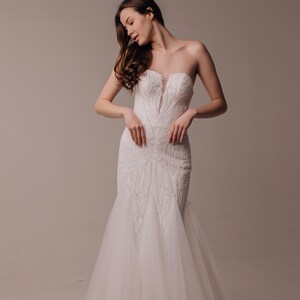 May include: A strapless white floor-length gown featuring a fitted bodice with intricate embroidery and beadwork, a deep plunging neckline with a sheer insert, and a flared skirt made of layered tulle that extends to the floor in a trumpet silhouette.