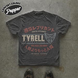 Peut inclure: Un t-shirt gris anthracite à manches courtes avec une impression graphique vieillie. Le motif comprend le texte "TYRELL CORPORATION", "GENETIC REPLICANTS", "MORE HUMAN THAN HUMAN", "LOS ANGELES CALIFORNIA" et des caractères japonais avec un logo hibou.