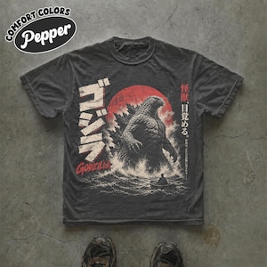 May include: A charcoal grey short-sleeve t-shirt featuring a graphic of Godzilla emerging from the ocean in front of a large red sun. The design includes Japanese text, the word Godzilla in red, and additional Japanese characters on the right side.