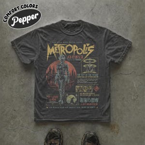 May include: A charcoal gray short-sleeve t-shirt featuring a graphic print inspired by the film Metropolis. The design includes the title in yellow, a central robot figure, red circular accents, and various text elements in English and Japanese.