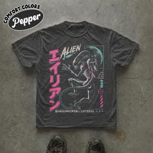 Peut inclure: Un t-shirt à manches courtes gris anthracite avec un motif d'Alien en bleu sarcelle et rose. Le design comprend le texte ALIEN, des caractères japonais et la phrase IN SPACE NO ONE CAN HEAR YOU SCREAM avec une icône de cible.