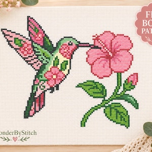 Peut inclure: Un modèle de point de croix sur tissu blanc représentant un colibri au plumage rose et vert près d'une grande fleur d'hibiscus rose. Le motif inclut un petit badge rose dans le coin supérieur droit avec le texte FREE BONUS PATTERN.