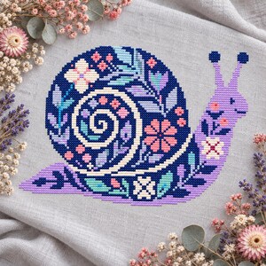 Può includere: Un ricamo a punto croce su tessuto grigio chiaro raffigurante una lumaca con un guscio blu scuro decorato con motivi floreali e di foglie rosa, azzurri e bianchi. Il corpo della lumaca è viola chiaro con piccoli accenti floreali.