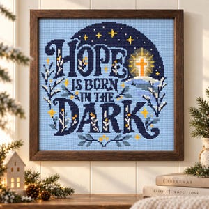 Könnte beinhalten: Ein quadratisches Kreuzstich-Stickbild in einem dunklen Holzrahmen mit dem Text HOPE IS BORN IN THE DARK in marineblauen Buchstaben. Das Design zeigt ein leuchtendes gelbes Kreuz, kleine gelbe Sterne und weiße florale Akzente auf hellblauem Grund.