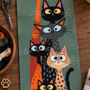 May include: A vertical cross-stitch embroidery on sage green fabric featuring a stack of six stylized cats. The cats vary in color, including orange, black, gray, and tan with spots, all depicted with large, wide eyes and long, thin bodies.
