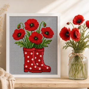 May include: A framed cross-stitch embroidery featuring a bouquet of vibrant red poppies emerging from a pair of red rain boots with white polka dots, set against a light gray grid background, displayed next to a glass vase of fresh red poppies.
