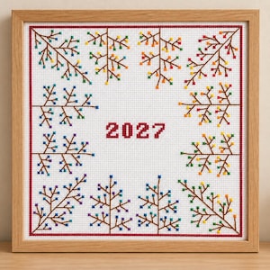 Può includere: Un ricamo a punto croce quadrato in una cornice di legno chiaro che mostra l'anno 2027 in rosso su tessuto bianco. Il disegno è bordato da una linea rossa e decorato con rami marroni con piccole foglie colorate a punto croce.