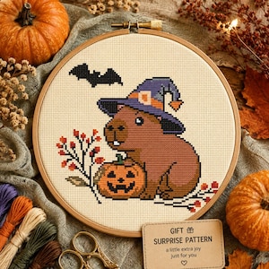 May include: A cross-stitch pattern in a wooden embroidery hoop featuring a brown capybara wearing a purple witch hat, a black bat, and a jack-o'-lantern pumpkin. The design includes a small tag with the text: GIFT SURPRISE PATTERN a little extra joy just for you.