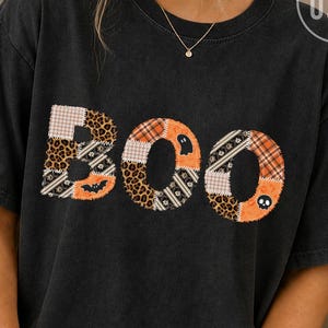 Op de afbeelding: Een zwart T-shirt met korte mouwen en het woord BOO in grote letters. Elke letter is samengesteld uit een patchworkontwerp met diverse patronen, waaronder luipaardprint, ruitjes en oranje stof met kleine spook- en schedelpictogrammen.