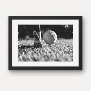Puede incluir: Una fotografía enmarcada en blanco y negro que muestra una pelota de golf sobre un tee junto a un palo de golf. La escena se sitúa sobre una superficie de césped, capturando los hoyuelos texturizados de la pelota y el acabado metálico del palo.