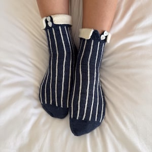 May include: A pair of navy blue knit socks featuring vertical white stripes and a folded cream-colored cuff. Each sock is accented with two small white heart-shaped buttons positioned on the outer side of the cuff, adding detail to the design.