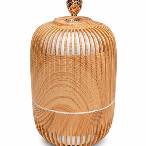 May include: A cylindrical essential oil diffuser with a light brown wood grain finish and vertical slats. The device features a white inner core and a metallic rose gold top accent piece. The structure is rounded with a visible horizontal seam.