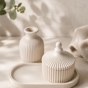 Könnte beinhalten: Ein cremefarbenes Set bestehend aus einem ovalen Tablett, einer kleinen Vase mit gerippter Textur und einem Dosendeckel mit passendem Rippendesign und einem abgerundeten Knauf. Die Teile bestehen aus einem festen, matten Material in hellem Beige.