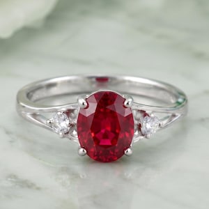 Può includere: Un anello color argento con una gemma centrale rosso intenso dal taglio ovale, fissata da quattro griffe. La fascia è divisa sulle spalle, con ogni lato impreziosito da una piccola pietra bianca rotonda incastonata nel gambo metallico.