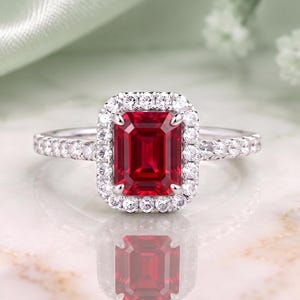 Pode incluir: Um anel de tom prateado com uma pedra preciosa vermelha retangular de corte esmeralda no centro, rodeada por um halo de pequenas pedras brancas redondas. A banda também é cravejada com uma série de pequenas pedras brancas redondas nos lados.
