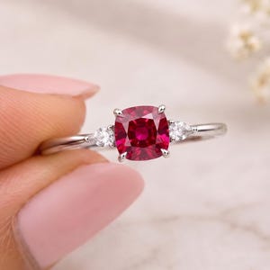 Könnte beinhalten: Ein silberfarbener Ring mit einem zentralen, kissenförmigen, tiefroten Edelstein, der von vier Krappen gehalten wird. Das Band ist auf jeder Seite mit einem kleinen, birnenförmigen, klaren Stein akzentuiert, was ein Drei-Stein-Design ergibt.