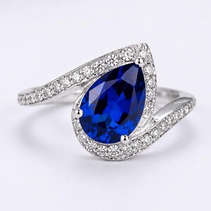 Könnte beinhalten: Ein silberfarbener Ring mit einem zentralen, birnenförmigen, tiefblauen Edelstein. Die Ringschiene und die Fassung um den birnenförmigen Stein sind mit kleinen, klaren, rund geschliffenen weißen Steinen besetzt, was ein Bypass-Design ergibt.