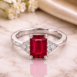 Puede incluir: Un anillo de plata con una piedra preciosa roja rectangular de corte esmeralda en el centro, flanqueada por dos grupos triangulares de pequeñas piedras blancas transparentes a cada lado, sobre una banda de plata pulida con acabado liso.