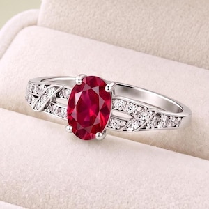 Può includere: Un anello color argento con una grande pietra preziosa rosso scuro dal taglio ovale al centro. La fascia è impreziosita da piccole pietre chiare dal taglio rotondo incastonate lungo i lati in un motivo metallico decorativo e traforato.