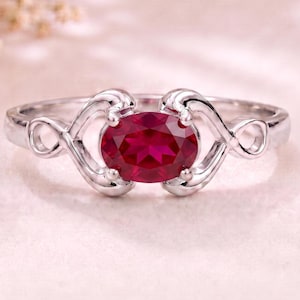Könnte beinhalten: Ein silberfarbener Ring mit einem zentralen, oval geschliffenen, tiefroten Edelstein. Die Ringschiene ist mit kunstvollen, geschwungenen Metallverzierungen gestaltet, die den Stein beidseitig einrahmen und ein fließendes Design bilden.