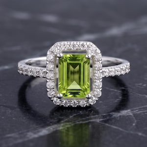 May include: A silver ring featuring a rectangular emerald-cut lime green peridot gemstone at the center. The stone is surrounded by a halo of small round white diamonds, with additional small white diamonds set along the thin silver band.
