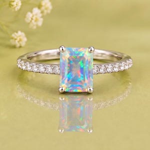 Può includere: Un anello color argento con una pietra centrale rettangolare in opale iridescente taglio smeraldo fissata da quattro griffe, con una fascia impreziosita da una fila di piccole pietre rotonde trasparenti su ciascun lato.