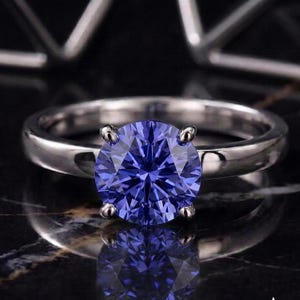 Peut inclure: Une bague argentée ornée d'une pierre précieuse bleue vibrante, taillée en brillant rond et fixée par quatre griffes. L'anneau est lisse et poli, reflétant la lumière, et la pierre présente une couleur bleue profonde et saturée avec des facettes.