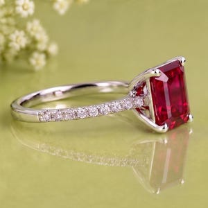 K&ouml;nnte beinhalten: Ein silberfarbener Ring mit einem gro&szlig;en, rechteckigen, tiefroten Edelstein im Smaragdschliff als Mittelstein. Die Ringschiene besteht aus poliertem Metall und ist mit einer Reihe kleiner, runder, klarer wei&szlig;er Steine besetzt.