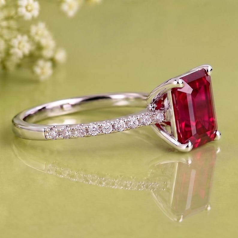 K&ouml;nnte beinhalten: Ein silberfarbener Ring mit einem gro&szlig;en, rechteckigen, tiefroten Edelstein im Smaragdschliff als Mittelstein. Die Ringschiene besteht aus poliertem Metall und ist mit einer Reihe kleiner, runder, klarer wei&szlig;er Steine besetzt.