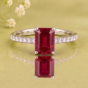 May include: A silver ring featuring a large, rectangular emerald-cut red gemstone as the centre stone. The band is set with a row of small, clear, round-cut stones along both sides, extending from the central setting to the back of the ring.