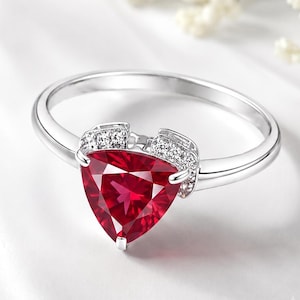 Pode incluir: Um anel de tom prateado com uma grande pedra preciosa vermelho-escura de corte triangular como peça central. A banda é acentuada com pequenas pedras claras de corte redondo cravadas no metal em ambos os lados da pedra vermelha central.