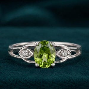 Op de afbeelding: Een zilverkleurige ring met een centraal ovaal geslepen limoengroene edelsteen in een zetting met vier pootjes. De band splitst zich aan weerszijden in twee armen, elk voorzien van een kleine, ronde, heldere steen in een marquisevormige zetting.