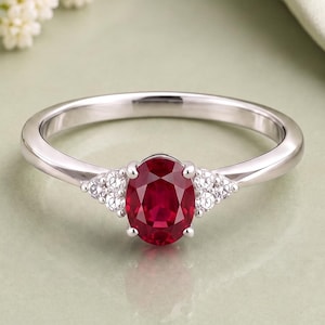 Könnte beinhalten: Ein silberfarbener Ring mit einem zentralen, oval geschliffenen, tiefroten Rubin. Der Rubin ist auf beiden Seiten von einer Gruppe aus drei kleinen, klaren, rund geschliffenen weißen Akzentsteinen flankiert, die in die polierte Ringschiene gefasst sind.