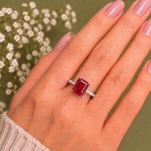K&ouml;nnte beinhalten: Ein silberfarbener Ring mit einem gro&szlig;en, rechteckigen, smaragdgeschliffenen roten Edelstein als Mittelstein. Die Ringschiene ist mit einer Reihe kleiner, klarer, rund geschliffener Steine besetzt, die f&uuml;r ein funkelndes Aussehen sorgen.