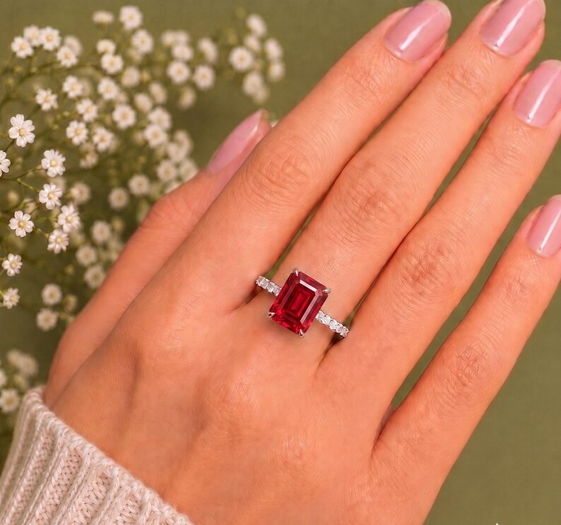 K&ouml;nnte beinhalten: Ein silberfarbener Ring mit einem gro&szlig;en, rechteckigen, smaragdgeschliffenen roten Edelstein als Mittelstein. Die Ringschiene ist mit einer Reihe kleiner, klarer, rund geschliffener Steine besetzt, die f&uuml;r ein funkelndes Aussehen sorgen.