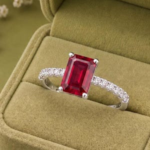 K&ouml;nnte beinhalten: Ein silberfarbener Ring mit einem gro&szlig;en, rechteckigen, tiefroten Edelstein im Smaragdschliff als Mittelstein, der von vier Krappen gehalten wird. Die Ringschiene ist mit einer Reihe kleiner, runder, klarer wei&szlig;er Steine besetzt.