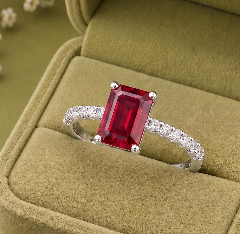 K&ouml;nnte beinhalten: Ein silberfarbener Ring mit einem gro&szlig;en, rechteckigen, tiefroten Edelstein im Smaragdschliff als Mittelstein, der von vier Krappen gehalten wird. Die Ringschiene ist mit einer Reihe kleiner, runder, klarer wei&szlig;er Steine besetzt.