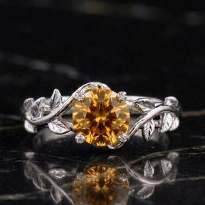 Puede incluir: Un anillo de tono plateado con una piedra preciosa redonda y facetada de color ámbar en el centro. La banda presenta un diseño detallado de enredaderas y hojas que envuelve la piedra central, asegurada por cuatro pequeñas garras metálicas.
