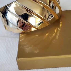 May include: A wide, polished gold-tone cuff bracelet featuring a series of diagonal, raised ridges that create a textured, reflective surface. The bracelet has a high-shine finish and a curved, structural design that wraps around the wrist.