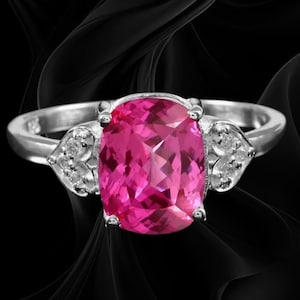 Puede incluir: Un anillo de plata con una gran piedra preciosa de color rosa vibrante y corte cojín como pieza central. La banda está acentuada a ambos lados por un engaste en forma de corazón que contiene tres pequeñas piedras claras y brillantes.