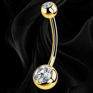 May include: A curved barbell belly button ring featuring a polished gold-tone metal finish. The piece includes a smaller top ball set with a single clear round gemstone and a larger bottom ball featuring a prominent, faceted clear round gemstone.
