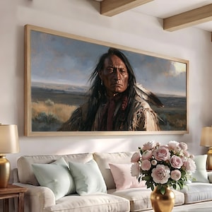 Puede incluir: Una impresión de arte mural rectangular en un marco de madera clara que muestra el retrato de una persona con cabello largo y oscuro y un accesorio de pluma, vistiendo una prenda beige con flecos, ante un paisaje vasto y apagado bajo un cielo nublado.
