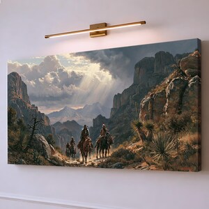 Può includere: Una stampa su tela rettangolare che raffigura un paesaggio montano desertico con tre figure a cavallo che percorrono un sentiero roccioso. La scena presenta nuvole drammatiche, raggi di sole, scogliere scoscese e vegetazione come yucca.