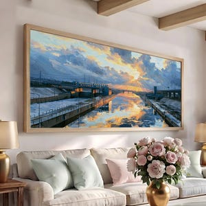 May include: A large rectangular landscape painting in a light wood frame depicting a sunset over a canal with industrial structures and bridges. The sky features thick clouds in shades of blue, orange, and yellow, reflecting on the water surface.