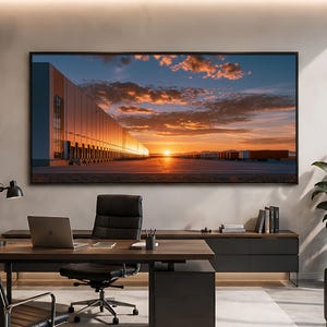 Puede incluir: Un gran cuadro rectangular de pared que muestra una puesta de sol sobre un largo almacén industrial con múltiples muelles de carga y remolques de camiones aparcados, en una oficina moderna con escritorio, silla y ordenador portátil.
