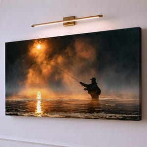 Könnte beinhalten: Ein rechteckiger Leinwanddruck, der die Silhouette einer Person beim Fliegenfischen auf einem nebligen See bei Sonnenaufgang zeigt. Die Szene besticht durch warmes orangefarbenes und goldenes Licht, das sich auf dem Wasser spiegelt, beleuchtet durch eine schmale, horizontale Messing-Wandleuchte.