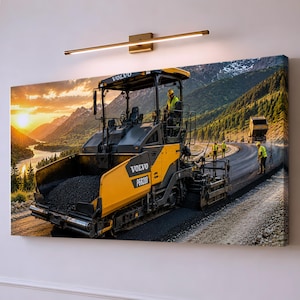 May include: A rectangular wall art print featuring a yellow and black Volvo P6820D asphalt paver on a road construction site. The scene includes workers in high-visibility vests, a dump truck, and a mountainous landscape at sunset.