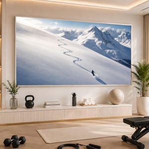 May include: A large rectangular wall-mounted television displaying a snowy mountain landscape with a skier leaving a winding track on a bright white slope. The screen is positioned above a long, light-colored media console with various home gym items.